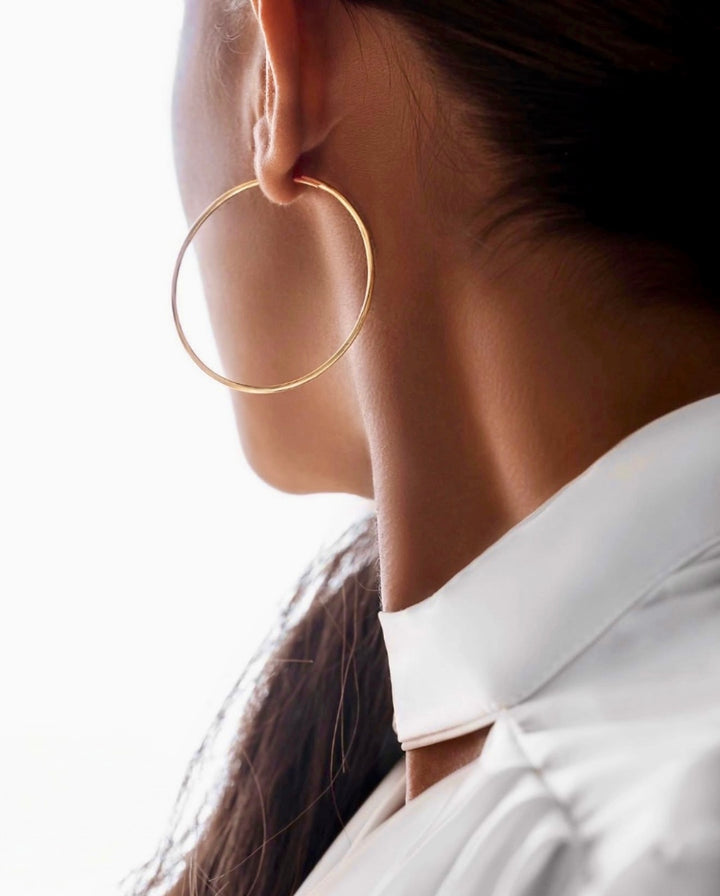 Elegante Nahaufnahme einer Person mit großem, dünnem Creolen-Ohrring in Gold. Das Haar ist zurückgebunden, der Fokus liegt auf Ohr, Hals und Schmuckstück. Die Person trägt ein weißes Oberteil mit hohem Kragen vor hellem, minimalistischem Hintergrund. Die Aufnahme wirkt modern, stilvoll und luxuriös.