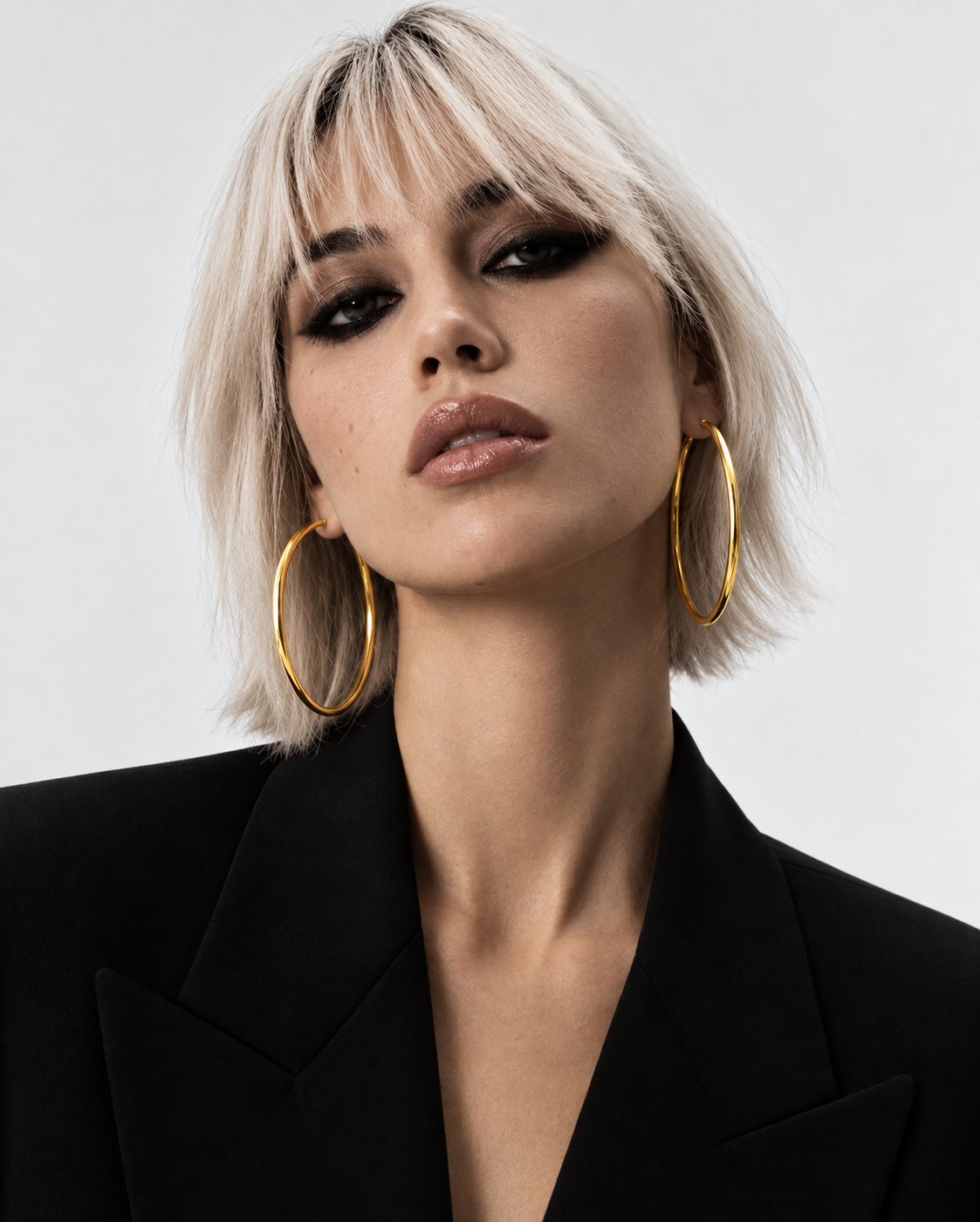 Fashion-Porträt einer Frau mit blondem Bob, schwarzem Blazer und grossen Creolen aus Gelbgold vor hellem Hintergrund.