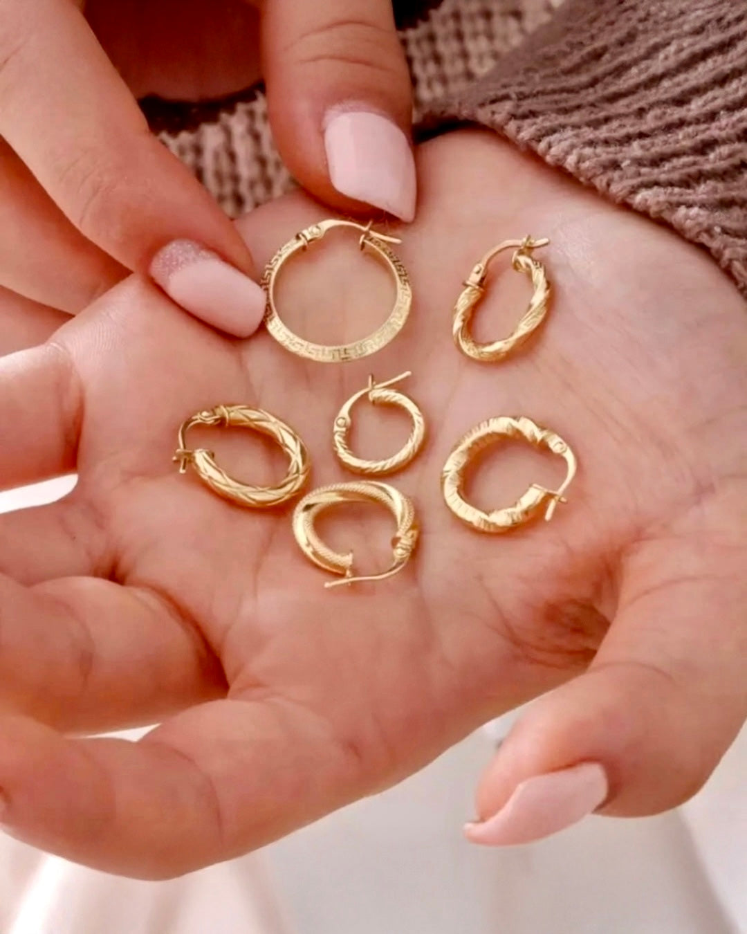 Hand mit sieben kleinen Creolen aus 18 Karat Gelbgold in unterschiedlichen Designs – glatt, gedreht, strukturiert und mit feinen Mustern – auf der offenen Handfläche präsentiert.