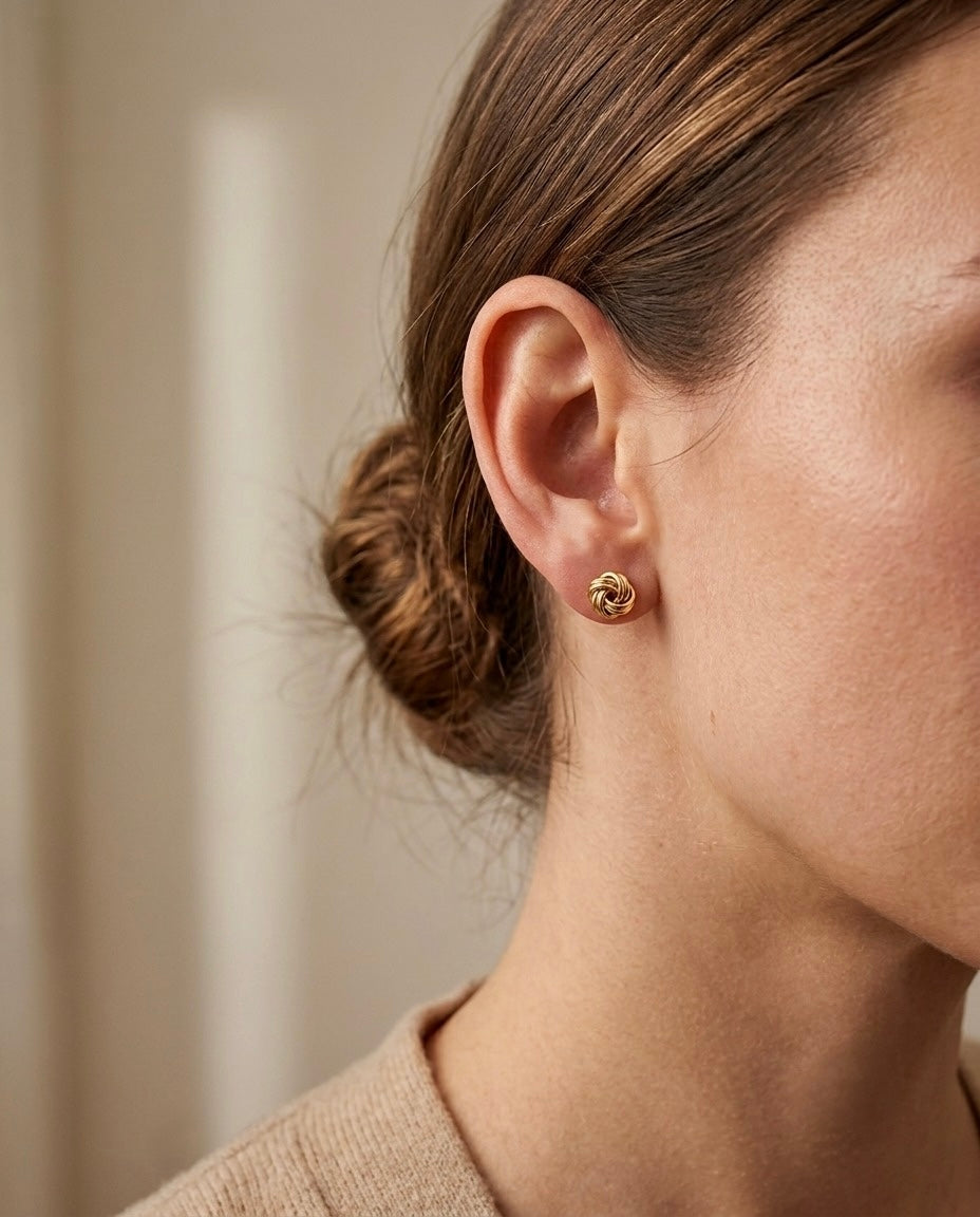 Elegante Nahaufnahme einer Person mit einem kleinen, runden Ohrstecker aus 18 Karat Gelbgold in gedrehter Knotenform. Das glänzend polierte Gold reflektiert das Licht dezent und verleiht dem zeitlosen Design eine edle Ausstrahlung. Der Ohrschmuck sitzt direkt am Ohrläppchen und passt sowohl zu eleganten als auch zu alltäglichen Outfits. Der weiche, unscharfe Hintergrund lenkt den Fokus auf den hochwertigen Goldohrring.
