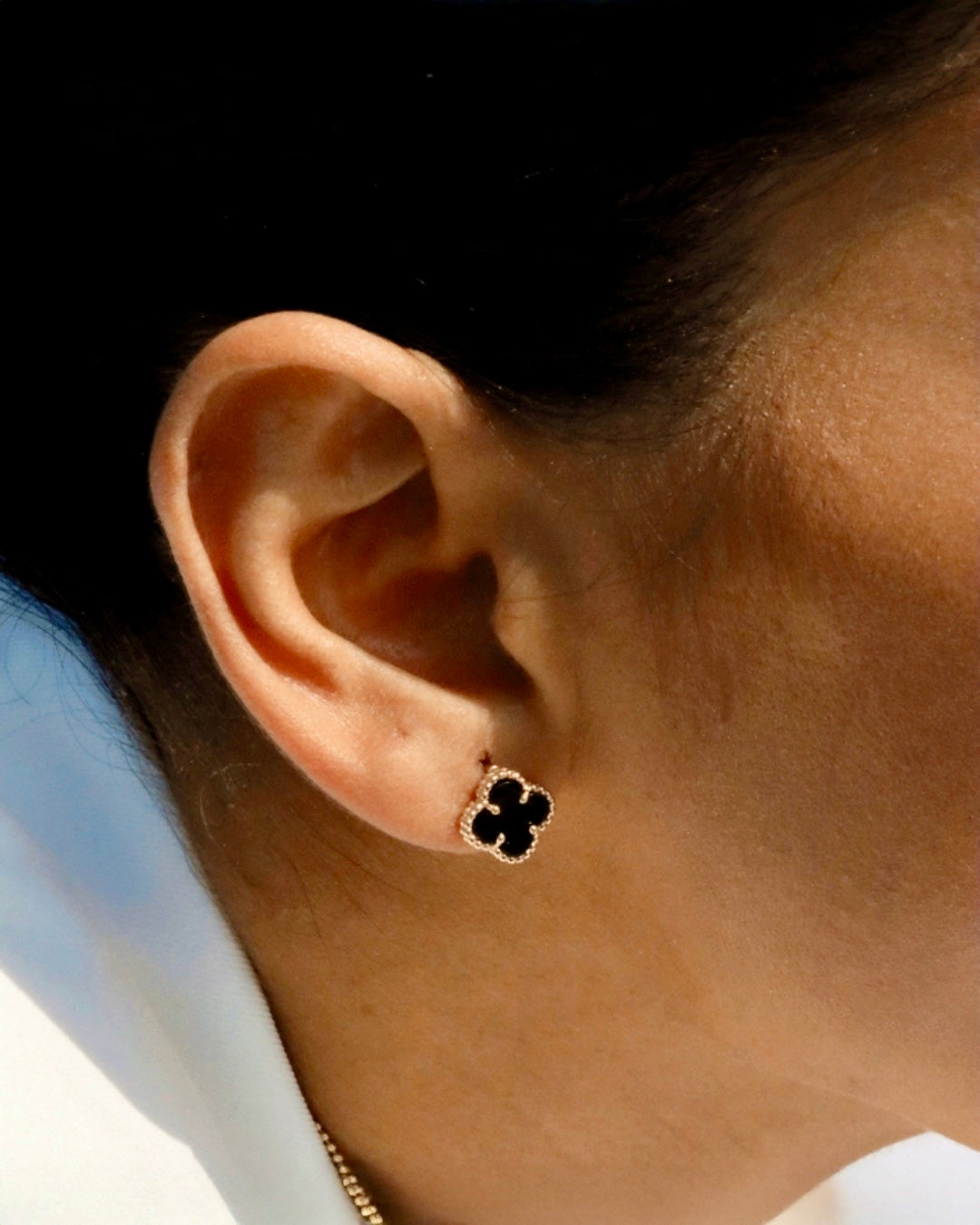 Nahaufnahme eines Ohres mit kleeblattförmigem Ohrstecker aus 18 Karat Gold mit schwarzem Onyx, elegant getragen vor neutralem Hintergrund.