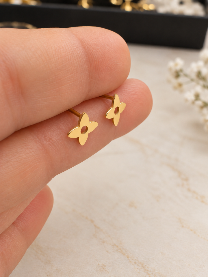Detailaufnahme zweier vierblättriger Goldohrstecker mit glatter Oberfläche und sichtbaren Steckstiften auf den Fingerspitzen.