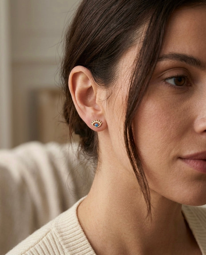 Frau trägt filigrane Evil-Eye-Ohrstecker aus 18 Karat Gelbgold mit blauem Nazar-Auge. Elegante, minimalistische Ohrstecker mit Schutzsymbol für den Alltag und besondere Anlässe.