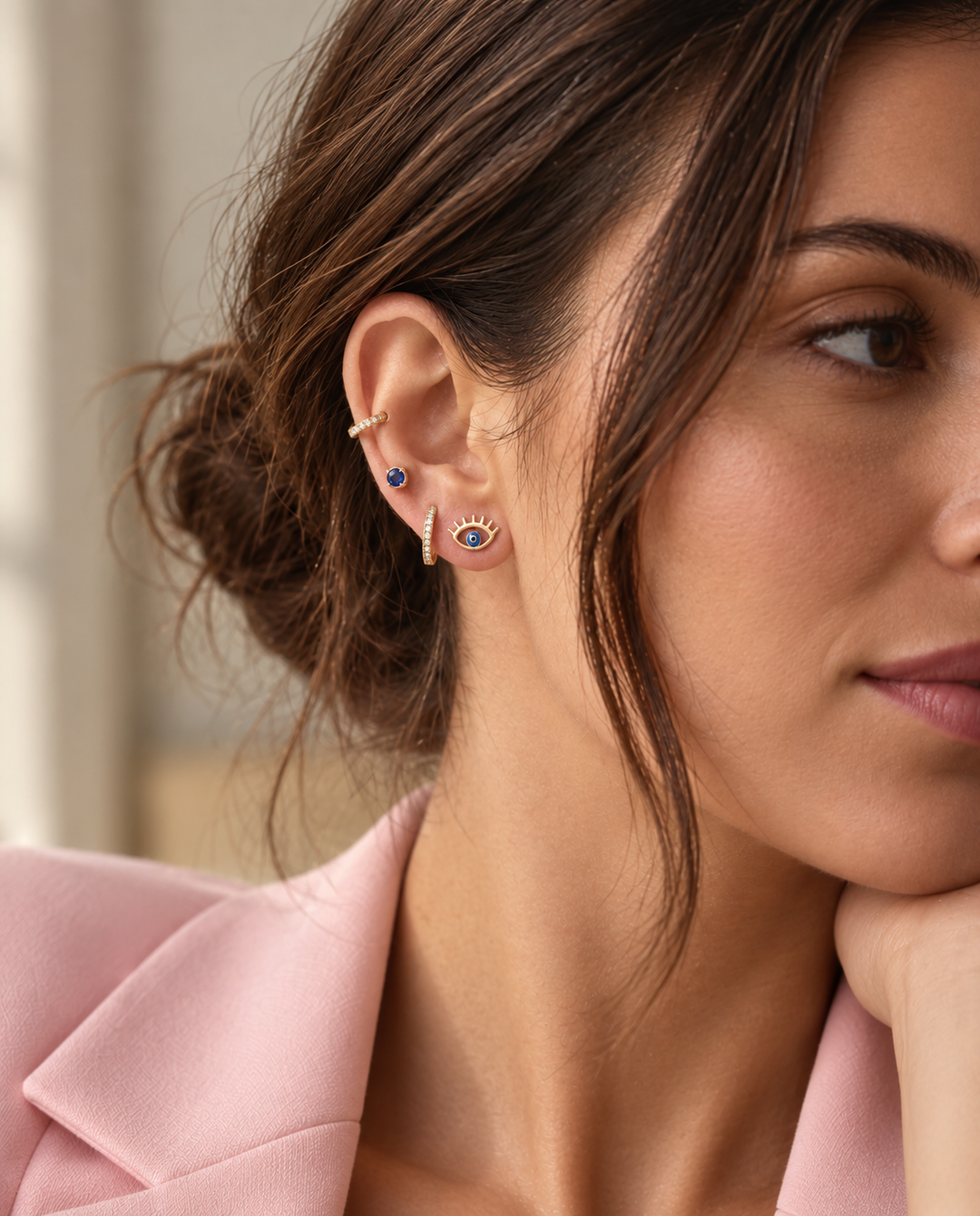 Frau mit rosafarbenem Blazer trägt ein Ear-Stacking aus goldenen Ohrringen mit blauem Augenmotiv, blauem Edelstein und funkelnden Creolen.