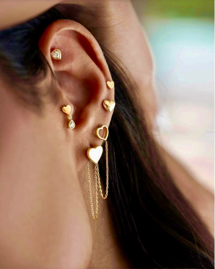 Detailaufnahme eines Ohrs mit mehreren feinen Ohrsteckern aus Gelbgold 18k in Herzform, teilweise mit kleinem Diamantbesatz und zarter Kettchen-Verbindung, elegant und modern kombiniert.