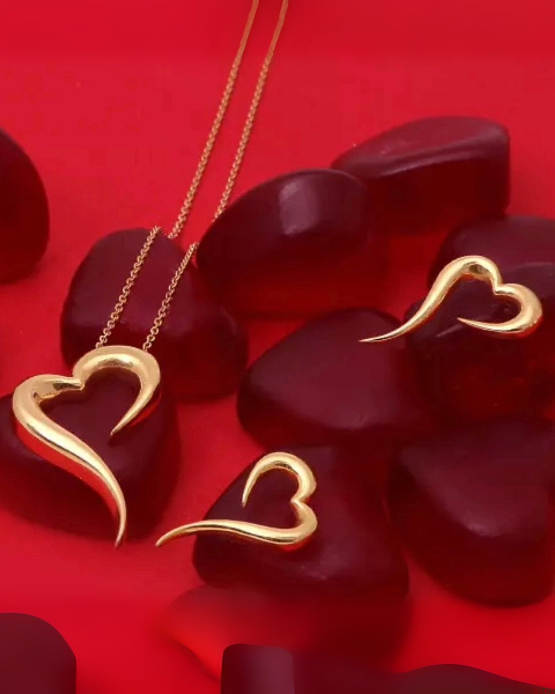 Goldschmuck-Set bestehend aus einer filigranen Halskette mit herzförmigem Anhänger und passenden, elegant geschwungenen Herz-Ohrringen, arrangiert auf roten, herzförmigen Elementen vor einem kräftig roten Hintergrund.