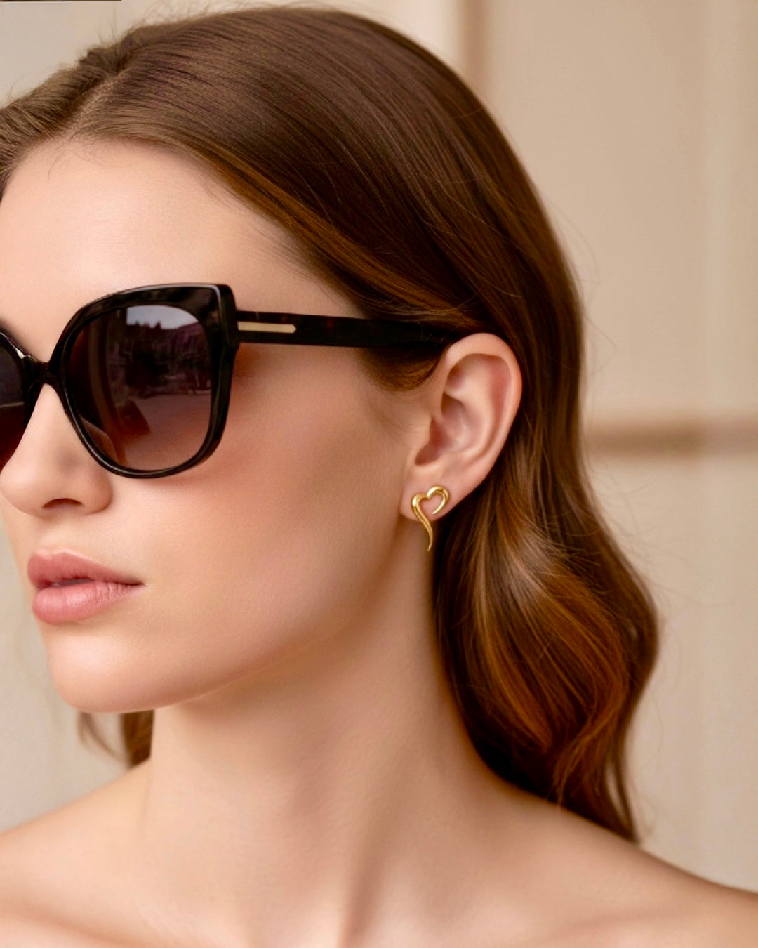 Nahaufnahme einer Frau mit glatter Haut und braunem Haar, die eine grosse schwarze Sonnenbrille und elegante, goldene, herzförmige Hängeohrringe trägt, vor einem weichen, neutralen Hintergrund.