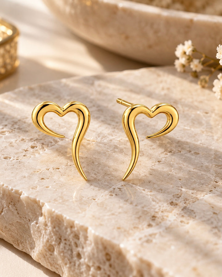 Goldene Ohrstecker in offener Herzform mit elegant geschwungenen Linien, inszeniert auf hellem Naturstein mit warmem Licht und zarten weissen Blüten.
