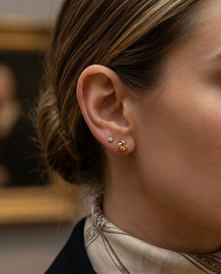 Frau trägt elegante 18-Karat-Gold-Ohrstecker in moderner Knotenform am Ohr. Die hochglanzpolierten Ohrstecker wirken zeitlos und stilvoll und lassen sich vielseitig zu Alltags- und Business-Outfits kombinieren. Ein kleiner Diamant-Ohrstecker im zweiten Ohrloch ergänzt den eleganten Look.