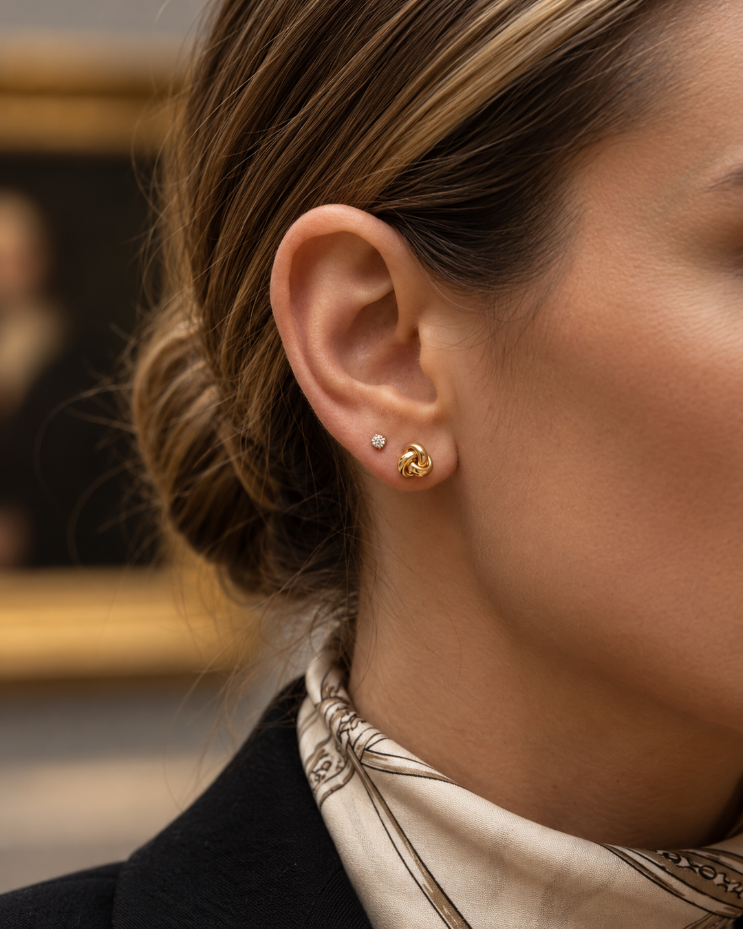 Frau trägt elegante 18-Karat-Gold-Ohrstecker in moderner Knotenform am Ohr. Die hochglanzpolierten Ohrstecker wirken zeitlos und stilvoll und lassen sich vielseitig zu Alltags- und Business-Outfits kombinieren. Ein kleiner Diamant-Ohrstecker im zweiten Ohrloch ergänzt den eleganten Look.