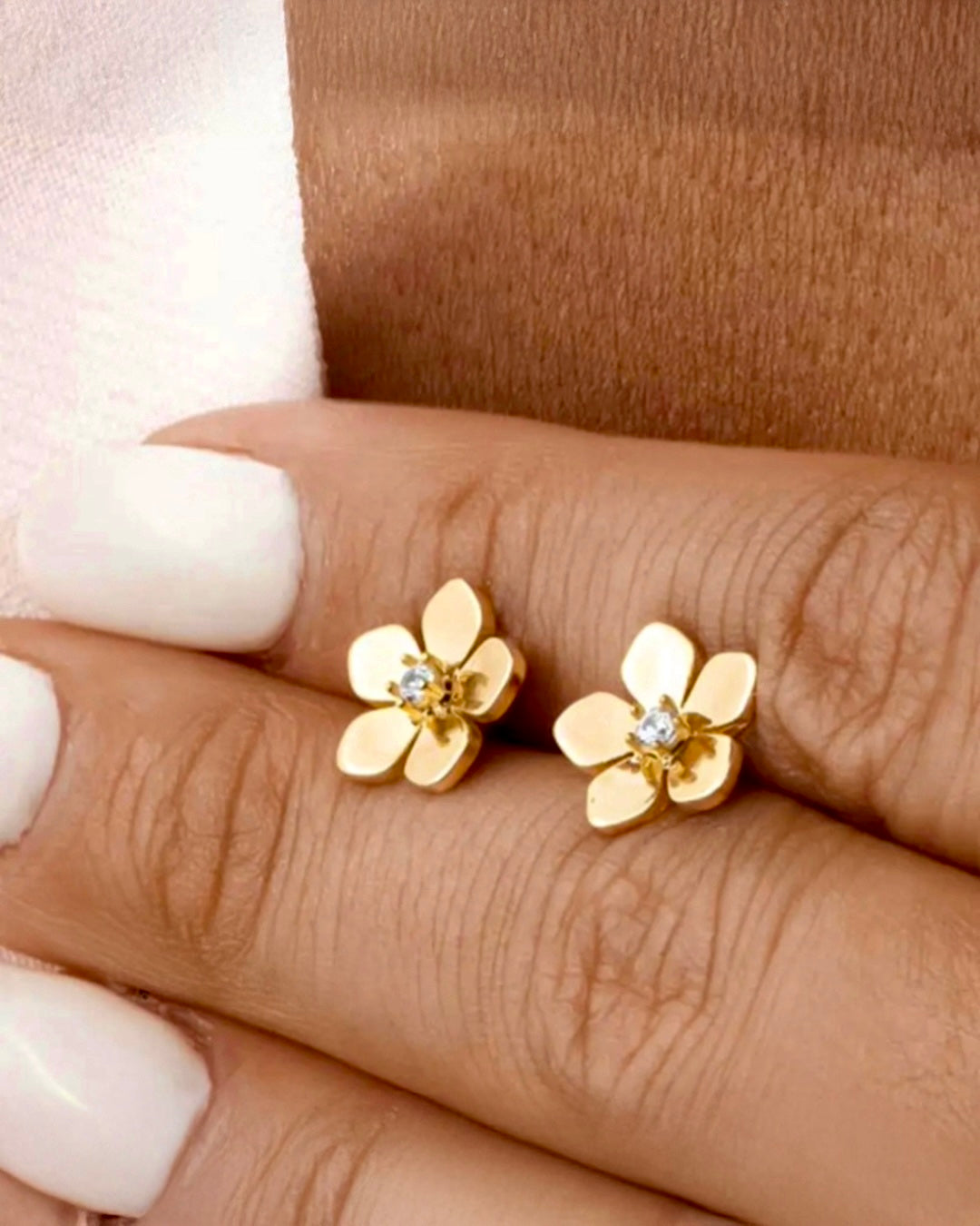 18k Gold-Blütenohrstecker, auf einer Hand zwischen zwei Fingern präsentiert – zarte Blütenform mit kleinem Stein in der Mitte.