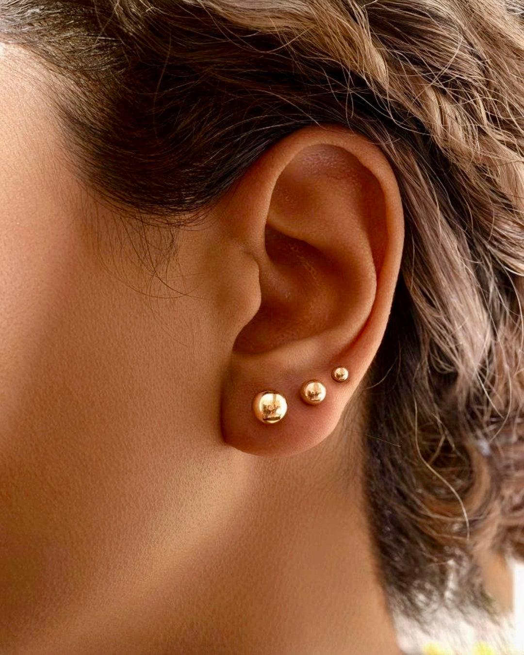 Nahaufnahme eines Ohres mit drei unterschiedlich grossen, runden Ohrsteckern aus poliertem 18-Karat-Gelbgold, dezent gestaffelt im Ohrloch getragen; minimalistischer Goldschmuck mit elegantem Glanz.