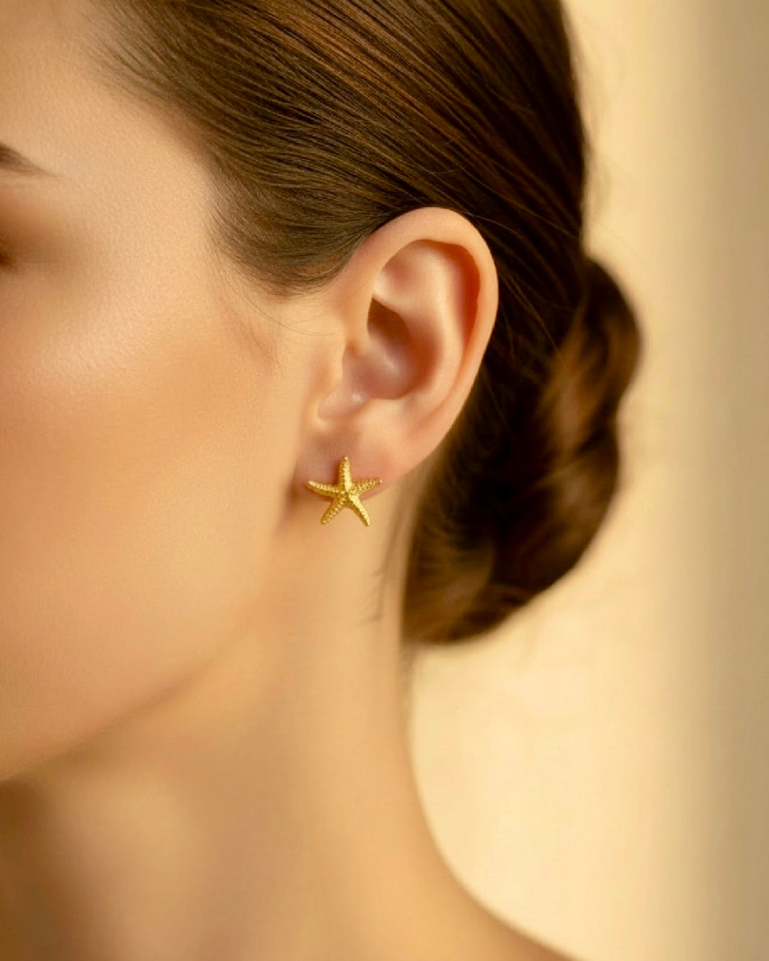 Goldene Seestern-Ohrstecker aus 18k Gelbgold am Ohr einer Frau, filigrane Struktur, elegantes Schmuckdetail im warmen Studio-Porträt.