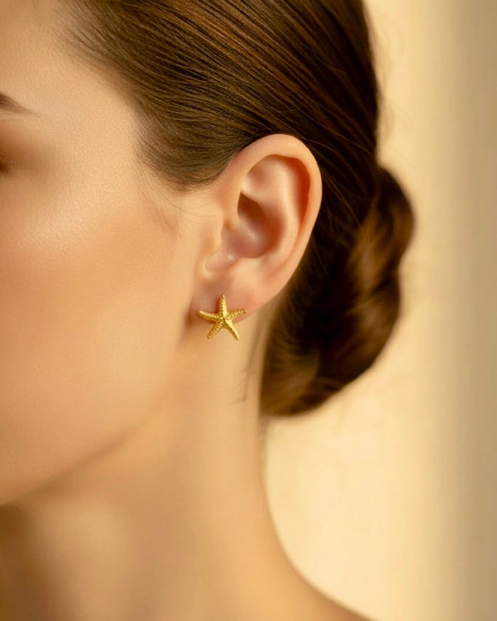 Goldene Seestern-Ohrstecker aus 18k Gelbgold am Ohr einer Frau, filigrane Struktur, elegantes Schmuckdetail im warmen Studio-Porträt.