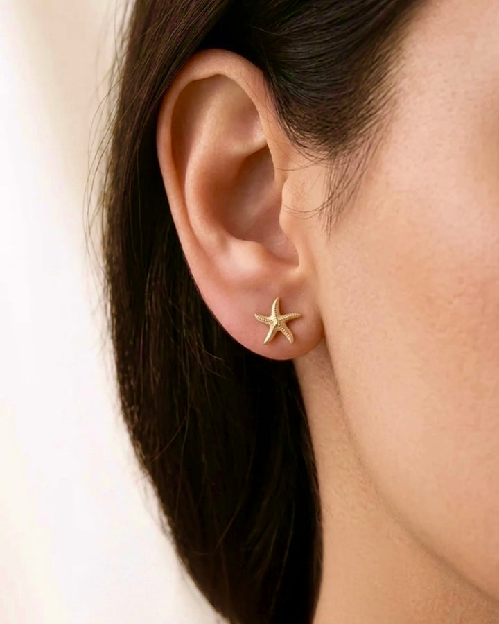 Nahaufnahme eines goldfarbenen Seestern-Ohrsteckers am Ohr einer Frau. Der filigrane Ohrschmuck im maritimen Design sitzt elegant am Ohrläppchen und wird vor einem weichen, neutralen Hintergrund präsentiert.