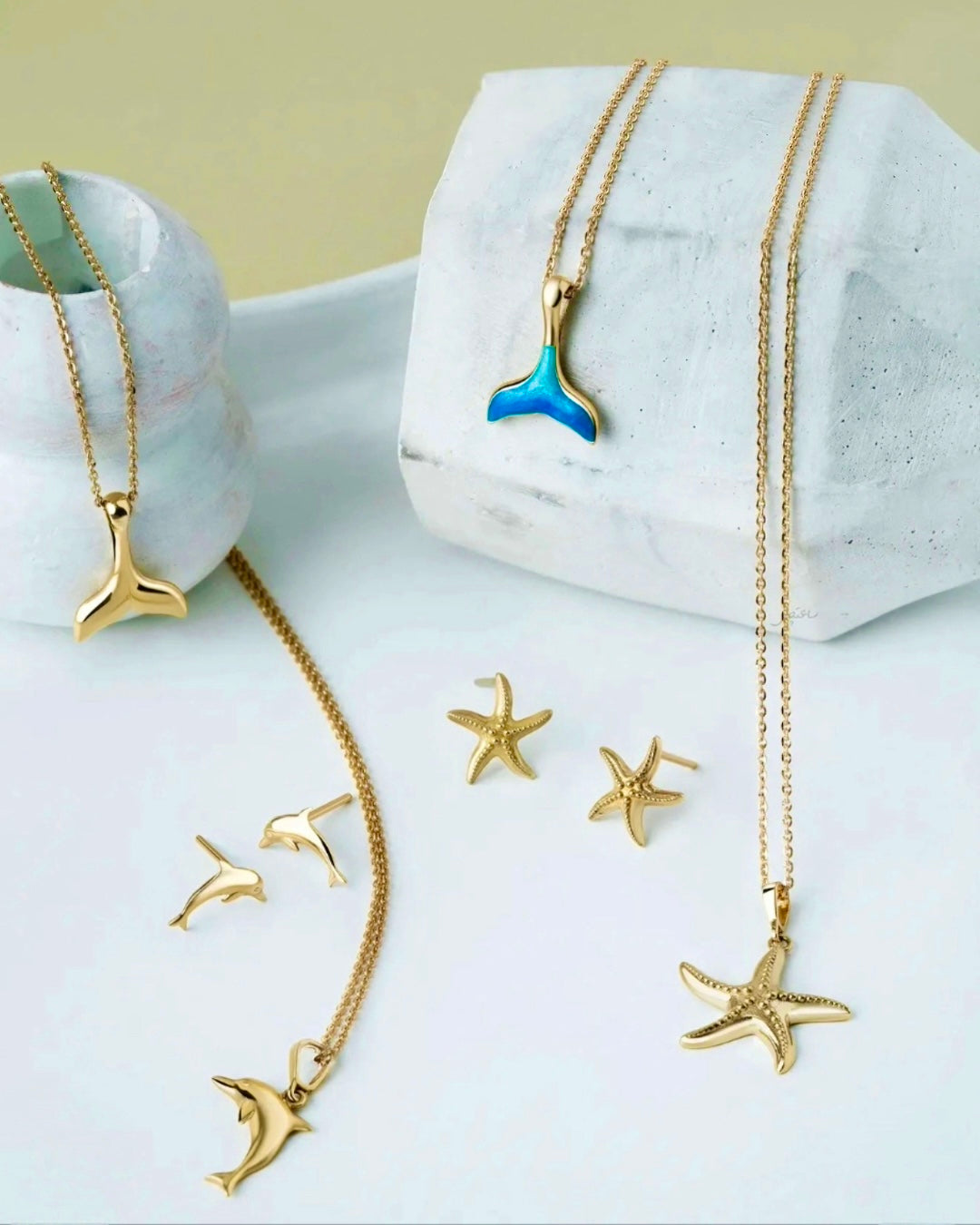 Elegante maritime Schmuckstücke aus goldfarbenem Edelstahl auf hellem Hintergrund: Halsketten mit Walflossen-, Seestern- und Delfin-Anhängern sowie passende Ohrringe mit Delfin- und Seestern-Motiven. Ein Anhänger zeigt eine blau-türkise Emaille-Walflosse. Minimalistische Präsentation mit weißen Dekoelementen im Hintergrund.