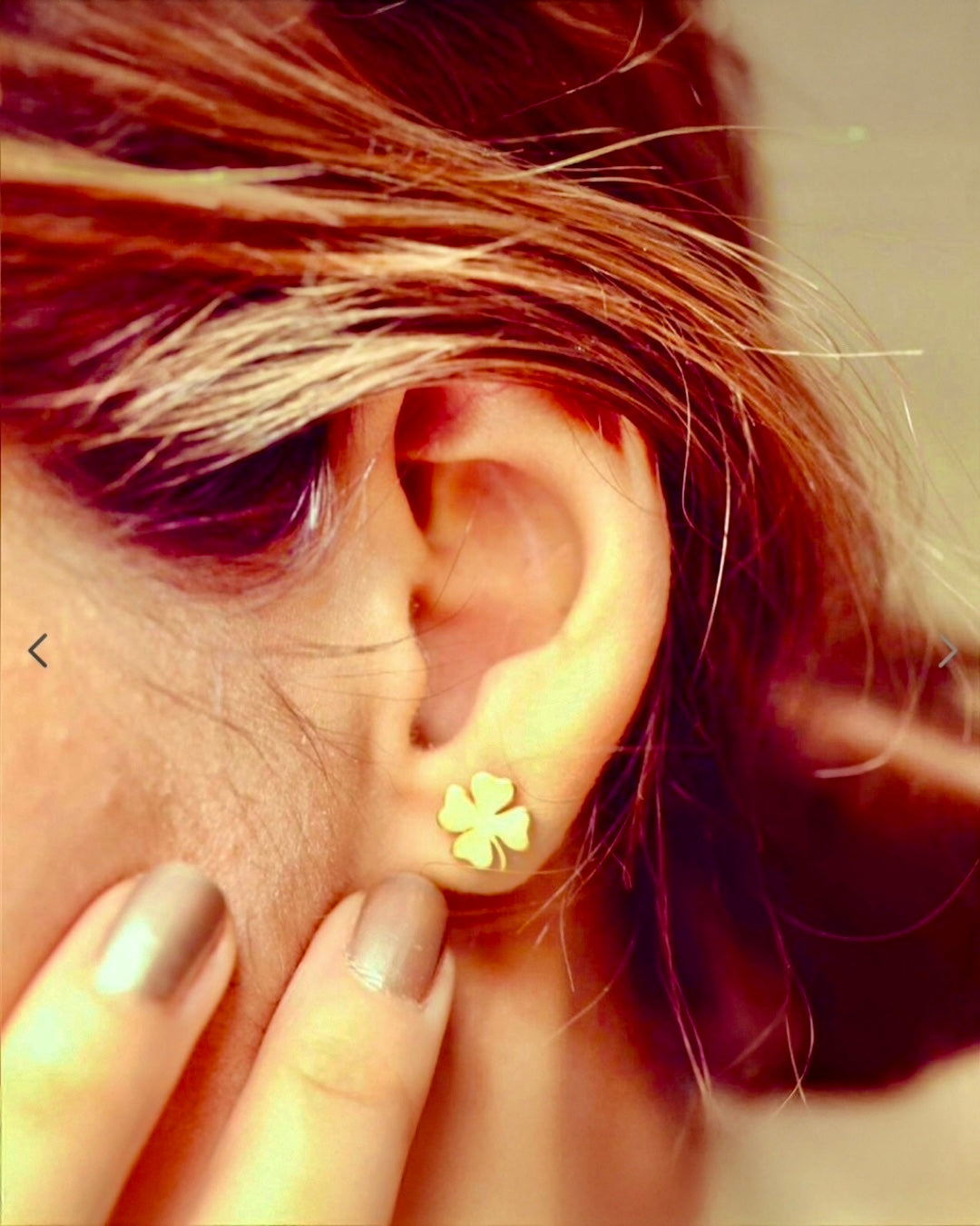 Nahaufnahme eines Ohrs mit einem kleinen, goldfarbenen Kleeblatt-Ohrstecker; eine Hand mit lackierten Fingernägeln berührt leicht die Wange.