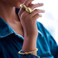 Elegante Nahaufnahme einer Frau mit modernem Goldschmuck: Zwei auffällige, skulpturale Goldringe an den Fingern und ein luxuriöses Goldarmband mit funkelnden Details am Handgelenk. Die Person trägt eine blaue Jeansjacke, während das natürliche Licht den hochglanzpolierten Schmuck edel hervorhebt.