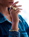 Elegante Nahaufnahme einer Frau mit modernem Goldschmuck: Zwei auffällige, skulpturale Goldringe an den Fingern und ein luxuriöses Goldarmband mit funkelnden Details am Handgelenk. Die Person trägt eine blaue Jeansjacke, während das natürliche Licht den hochglanzpolierten Schmuck edel hervorhebt.
