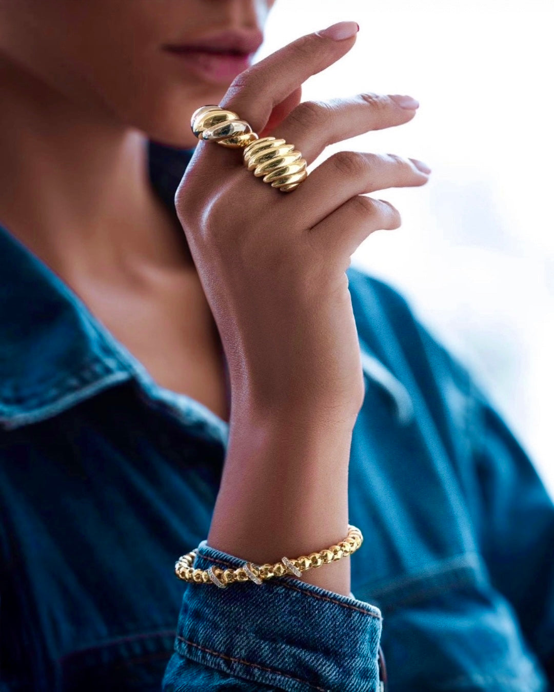 Elegante Nahaufnahme einer Frau mit modernem Goldschmuck: Zwei auffällige, skulpturale Goldringe an den Fingern und ein luxuriöses Goldarmband mit funkelnden Details am Handgelenk. Die Person trägt eine blaue Jeansjacke, während das natürliche Licht den hochglanzpolierten Schmuck edel hervorhebt.