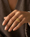 Nahaufnahme einer Hand mit gepflegten, naturfarben lackierten Fingernägeln. Am Ringfinger wird ein kunstvoll gefertigter Ring aus 18 Karat Gelbgold getragen. Das filigrane Design bildet eine elegante Unendlichkeitsform (∞) mit durchbrochener Struktur und diamantierter Oberfläche, die das Licht dezent reflektiert. Der Ring wirkt zugleich modern, feminin und zeitlos und wird vor einem warmen, weichen Hintergrund stilvoll präsentiert.
