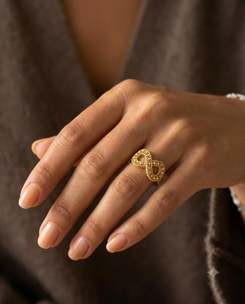 Nahaufnahme einer Hand mit gepflegten, naturfarben lackierten Fingernägeln. Am Ringfinger wird ein kunstvoll gefertigter Ring aus 18 Karat Gelbgold getragen. Das filigrane Design bildet eine elegante Unendlichkeitsform (∞) mit durchbrochener Struktur und diamantierter Oberfläche, die das Licht dezent reflektiert. Der Ring wirkt zugleich modern, feminin und zeitlos und wird vor einem warmen, weichen Hintergrund stilvoll präsentiert.