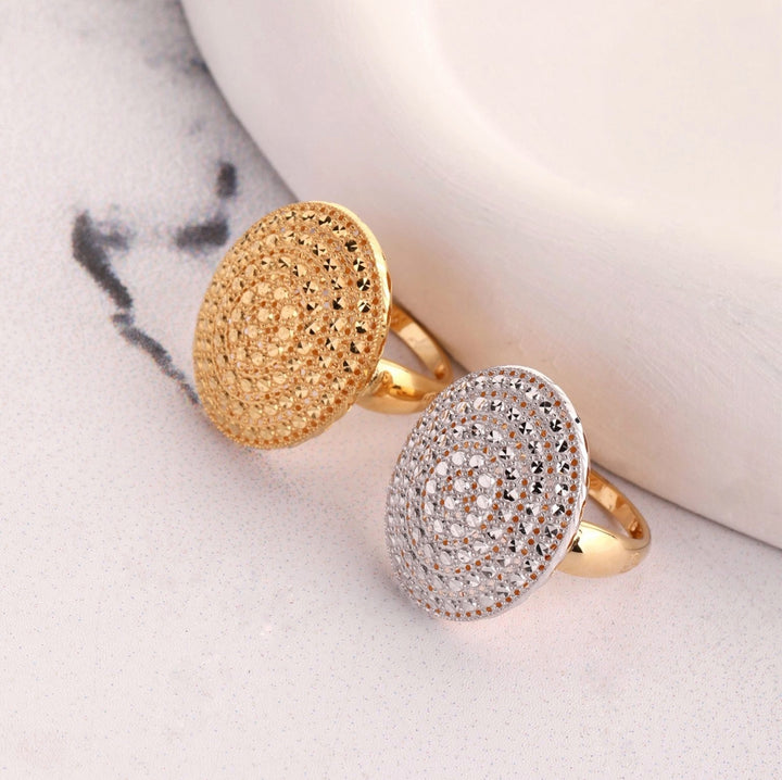 Zwei 18k Gelbgold-Ringe mit großem, rundem, filigran durchbrochenem Sonnenscheiben-Design, einer vollständig in Gelbgold, der andere mit rhodiniertem Weißgold-Akzent, auf heller Steinoberfläche arrangiert.