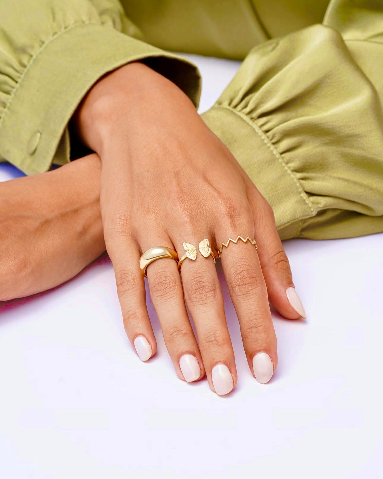Hand mit drei Ringen aus 18 Karat Gelbgold, darunter der Ring „Baleh tâ Hamisheh“ – filigranes Design mit zwei Schmetterlingsflügeln als Symbol für ein „Ja für immer“, elegant am Finger getragen.