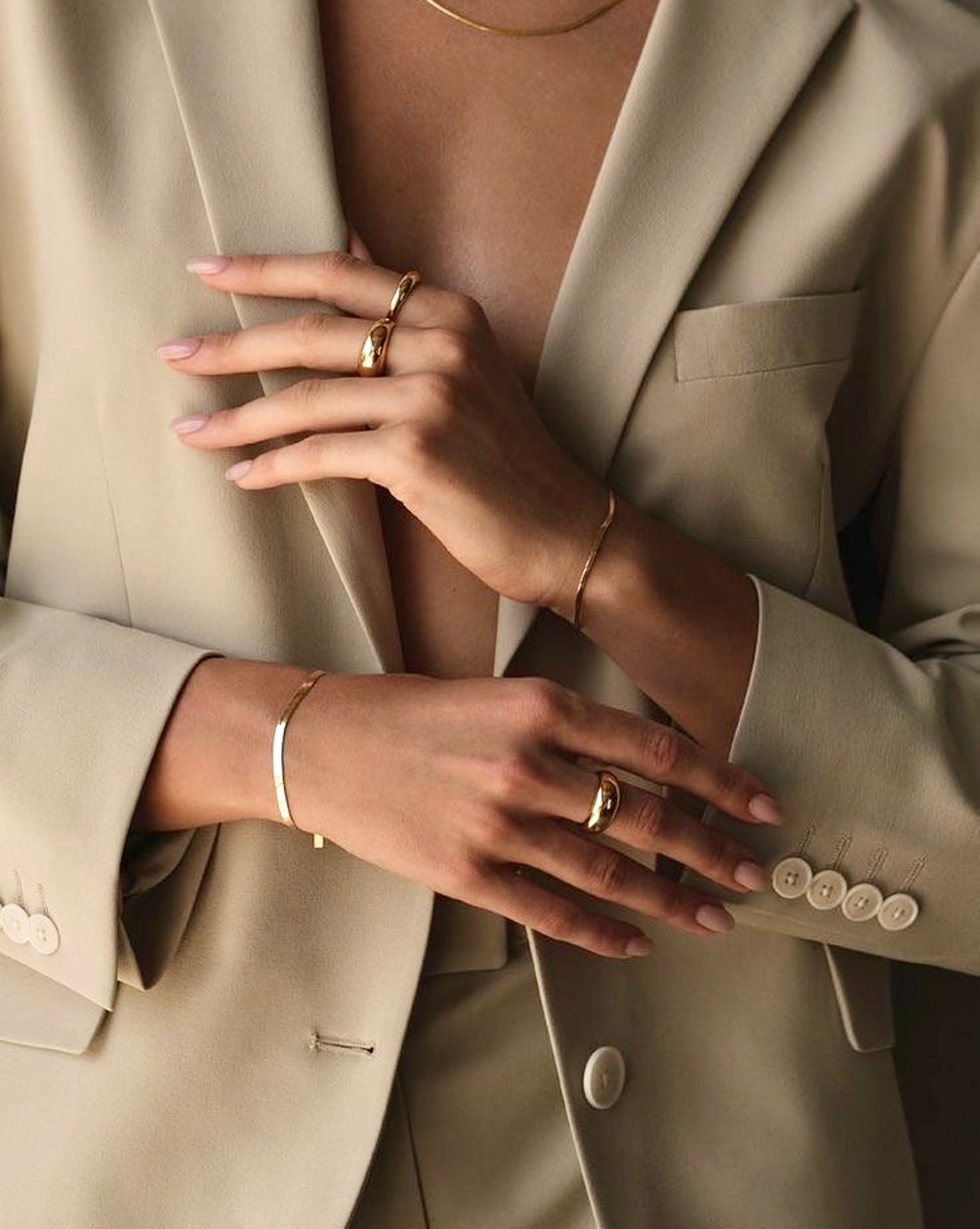 Elegantes Detailfoto einer Frau in hellem Blazer, die mehrere schlichte Ringe, feine Armreifen und eine zarte Halskette aus 18-karätigem Gelbgold trägt – minimalistisch, modern und stilvoll inszeniert.
