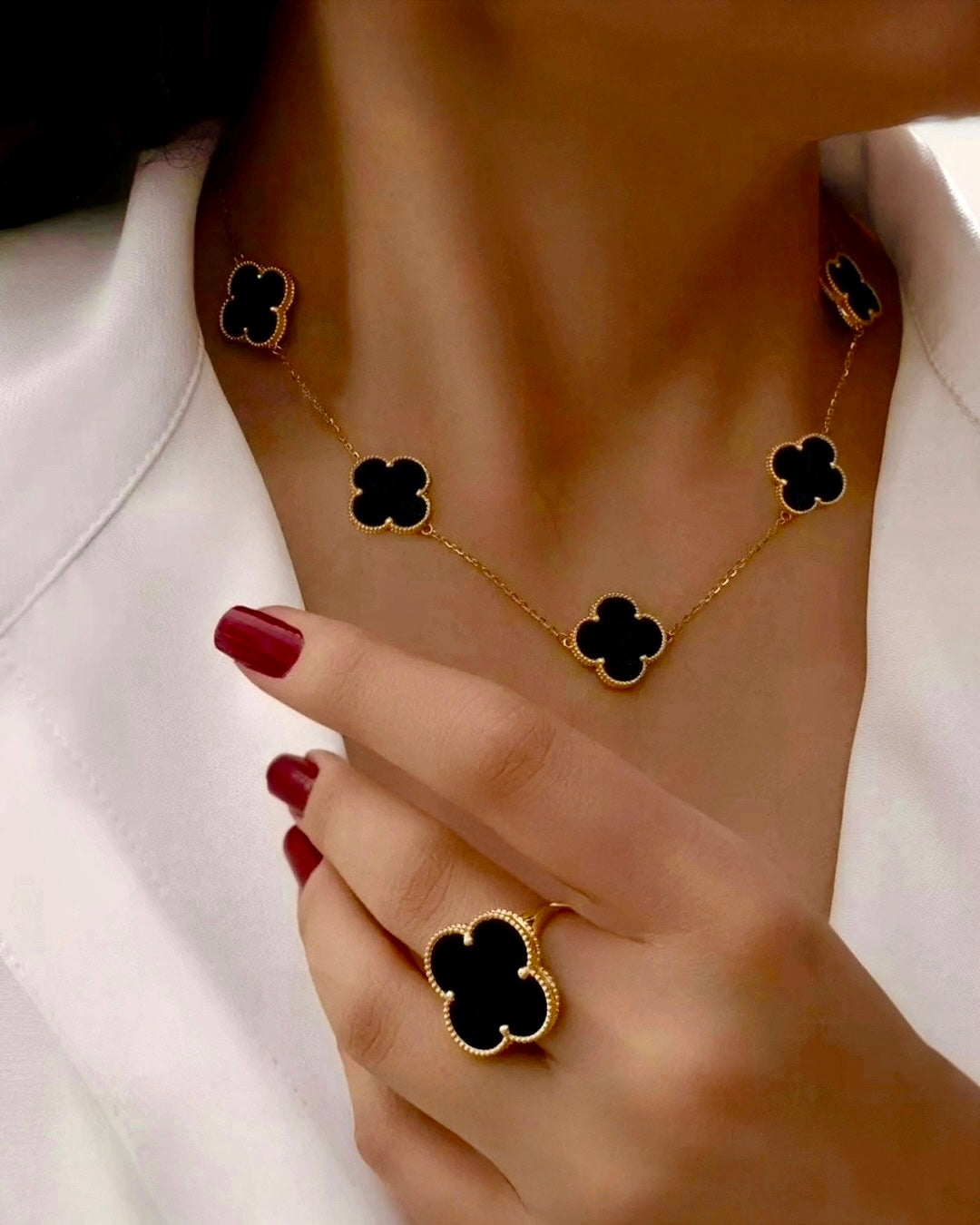 Nahaufnahme einer Frau mit einer Halskette und einem Ring aus 18 Karat Gelbgold – beide Schmuckstücke mit schwarzem Onyx in Kleeblattform („Barg-e Onyx“) gefasst, elegant kombiniert mit rotem Nagellack und weißem Outfit.