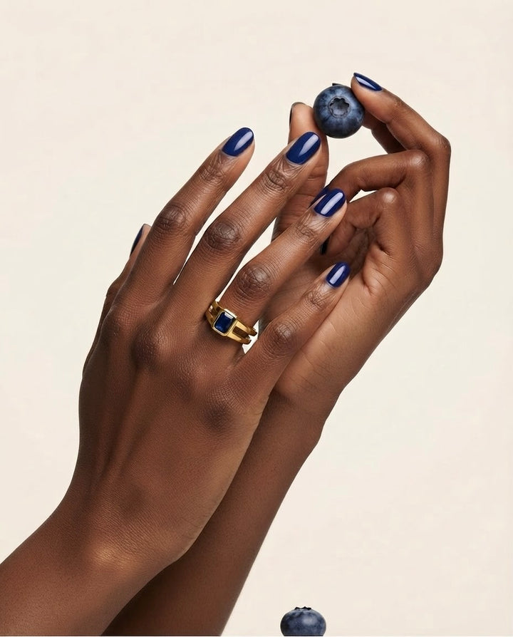Nahaufnahme zweier Hände mit dunkelblau lackierten Nägeln. Eine Hand trägt einen goldenen Ring mit rechteckigem dunkelblauem Stein, während die andere eine Heidelbeere zwischen den Fingern hält. Heller, minimalistischer Hintergrund.