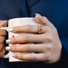Hand hält eine Tasse, geschmückt mit dem Ring „Golbarg-e Pichideh“ aus 18 Karat Gelbgold – detailreich verflochtenes Blütenmuster, kunstvoll gearbeitet und elegant im Alltag tragbar.