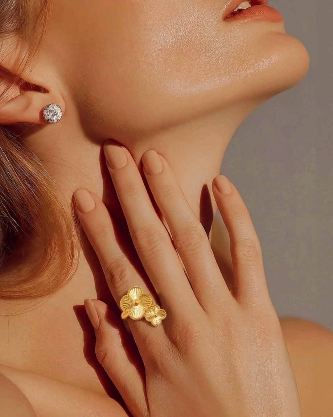Nahaufnahme von Hals und unterem Gesicht einer Frau im warmen Licht, mit einem funkelnden Ohrstecker. Ihre Hand liegt sanft am Hals und präsentiert einen goldenen Ring mit zwei filigranen Blumenmotiven.