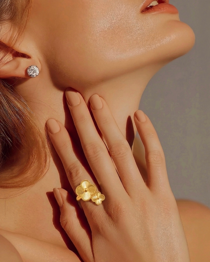 Nahaufnahme von Hals und unterem Gesicht einer Frau im warmen Licht, mit einem funkelnden Ohrstecker. Ihre Hand liegt sanft am Hals und präsentiert einen goldenen Ring mit zwei filigranen Blumenmotiven.