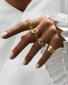 Hand mit mehreren modernen, massiven Goldringen in unterschiedlichen Designs, kombiniert mit weisser Bluse – eleganter Goldschmuck im Layering-Look.