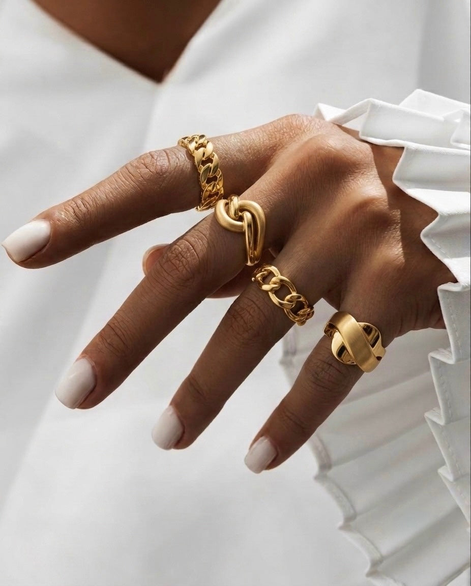 Hand mit mehreren modernen, massiven Goldringen in unterschiedlichen Designs, kombiniert mit weisser Bluse – eleganter Goldschmuck im Layering-Look.