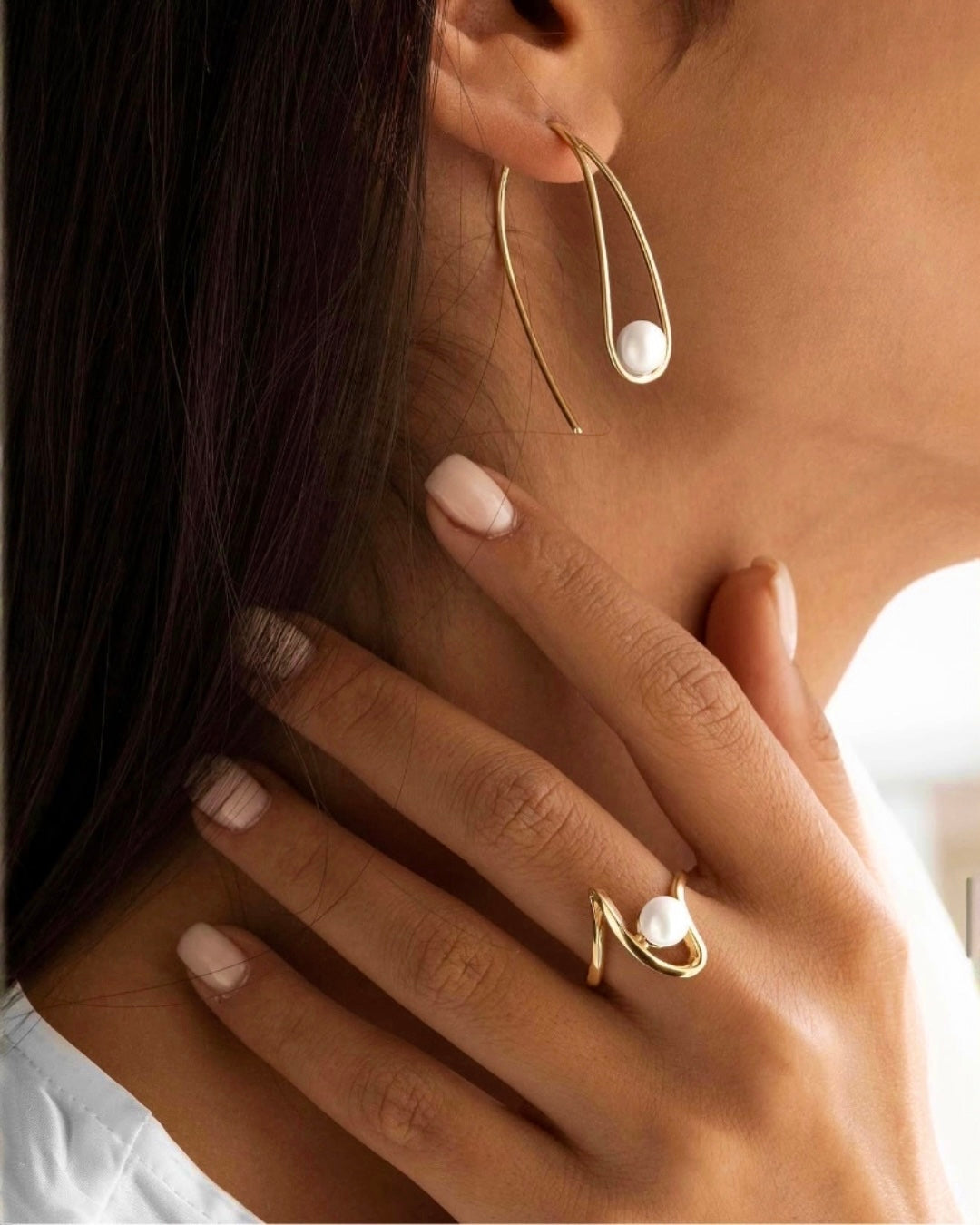 Elegante Detailaufnahme einer Frau mit modernem Goldschmuck: große tropfenförmige Creolen mit weißer Perle sowie passender offener Ring in geschwungenem Design mit Perle. Die warmen Goldtöne harmonieren mit der natürlichen Maniküre und verleihen dem Look eine stilvolle, minimalistische Eleganz.