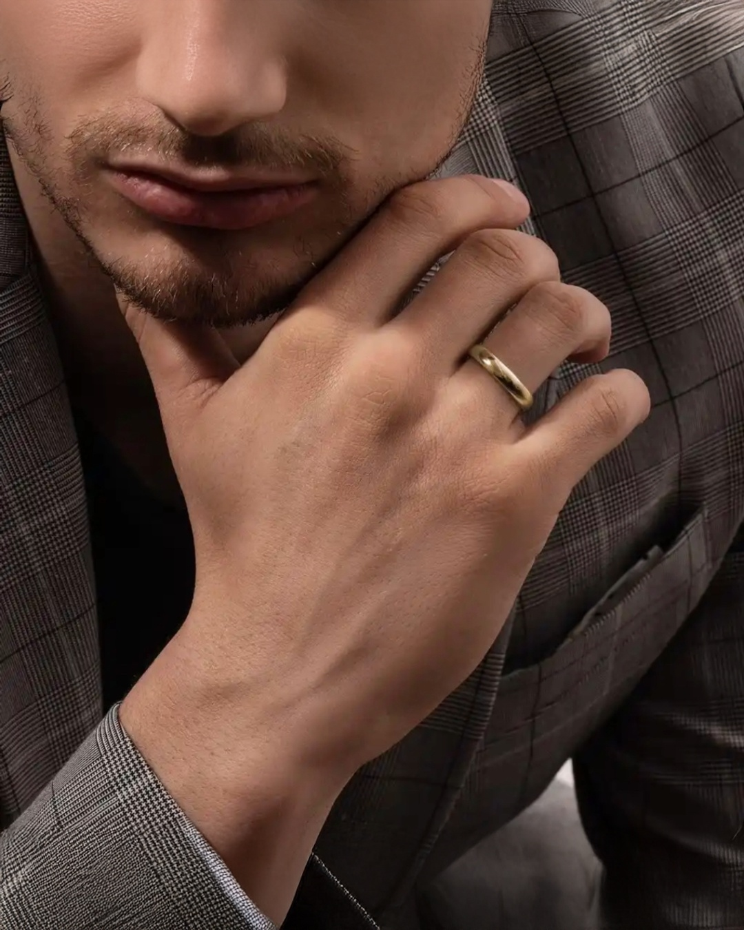Detailaufnahme eines Mannes im grauen Anzug, der nachdenklich seine Hand ans Kinn legt und einen schlichten, polierten Ring aus 18k Gelbgold am Ringfinger trägt.