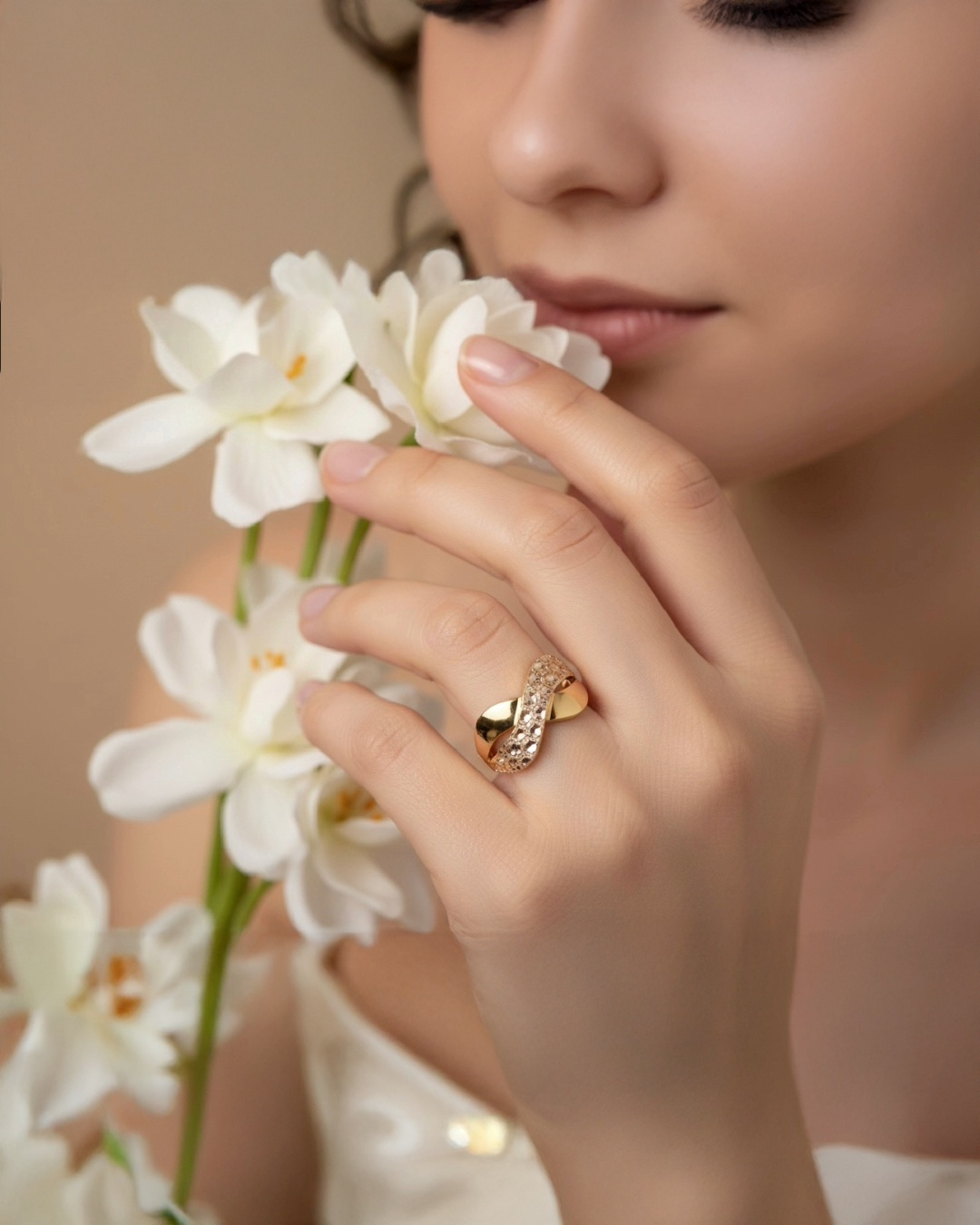 Frau mit eleganter 18k-Gelbgold-Ringkreation mit ineinander verschlungenem Design und funkelnden Akzenten, getragen an der Hand, sanft inszeniert mit weißen Blüten vor warmem, weichem Hintergrund.
