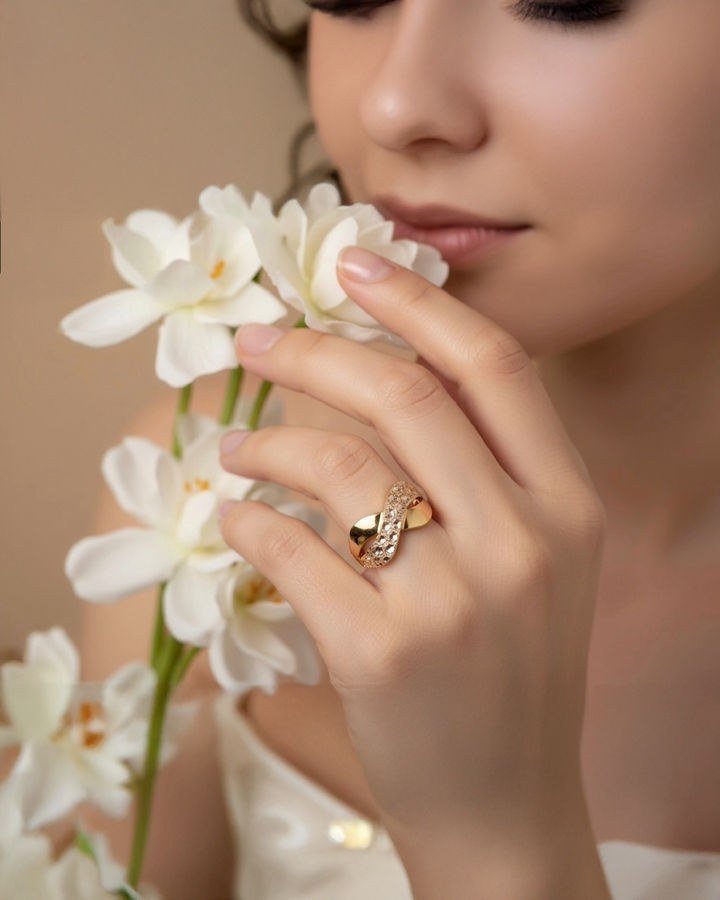 Frau mit eleganter 18k-Gelbgold-Ringkreation mit ineinander verschlungenem Design und funkelnden Akzenten, getragen an der Hand, sanft inszeniert mit weißen Blüten vor warmem, weichem Hintergrund.