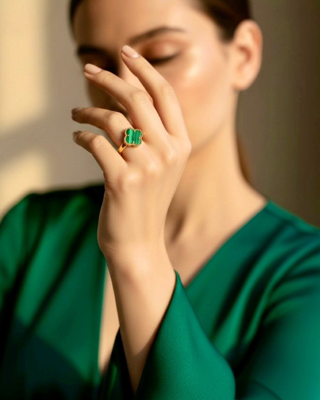 Elegant woman wearing a deep green dress, gently touching her face while showcasing a refined 18k gold ring with a clover-shaped green gemstone (malachite style) set in a delicate gold frame; soft lighting highlights the ring’s rich color and luxurious finish.