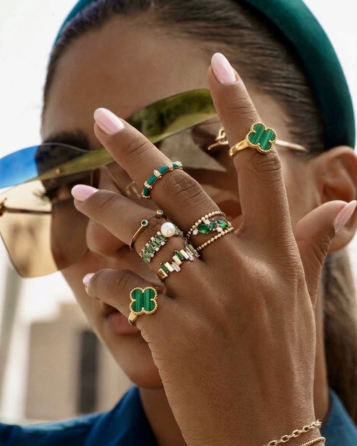 Elegante Nahaufnahme einer Frau mit Sonnenbrille, die mehrere luxuriöse Ringe in Grün-, Gold- und Weissgoldtönen trägt. Die Hand steht im Fokus und präsentiert verschiedene Schmuckstücke mit grünen Edelsteinen, Perlen und geometrischen Designs. Besonders auffällig sind zwei vierblättrige Kleeblatt-Ringe aus grünem Malachit mit goldener Fassung. Die Kombination aus modernen Accessoires, gepflegten rosafarbenen Nägeln und edlem Schmuck vermittelt einen exklusiven, stilvollen Luxus-Look.