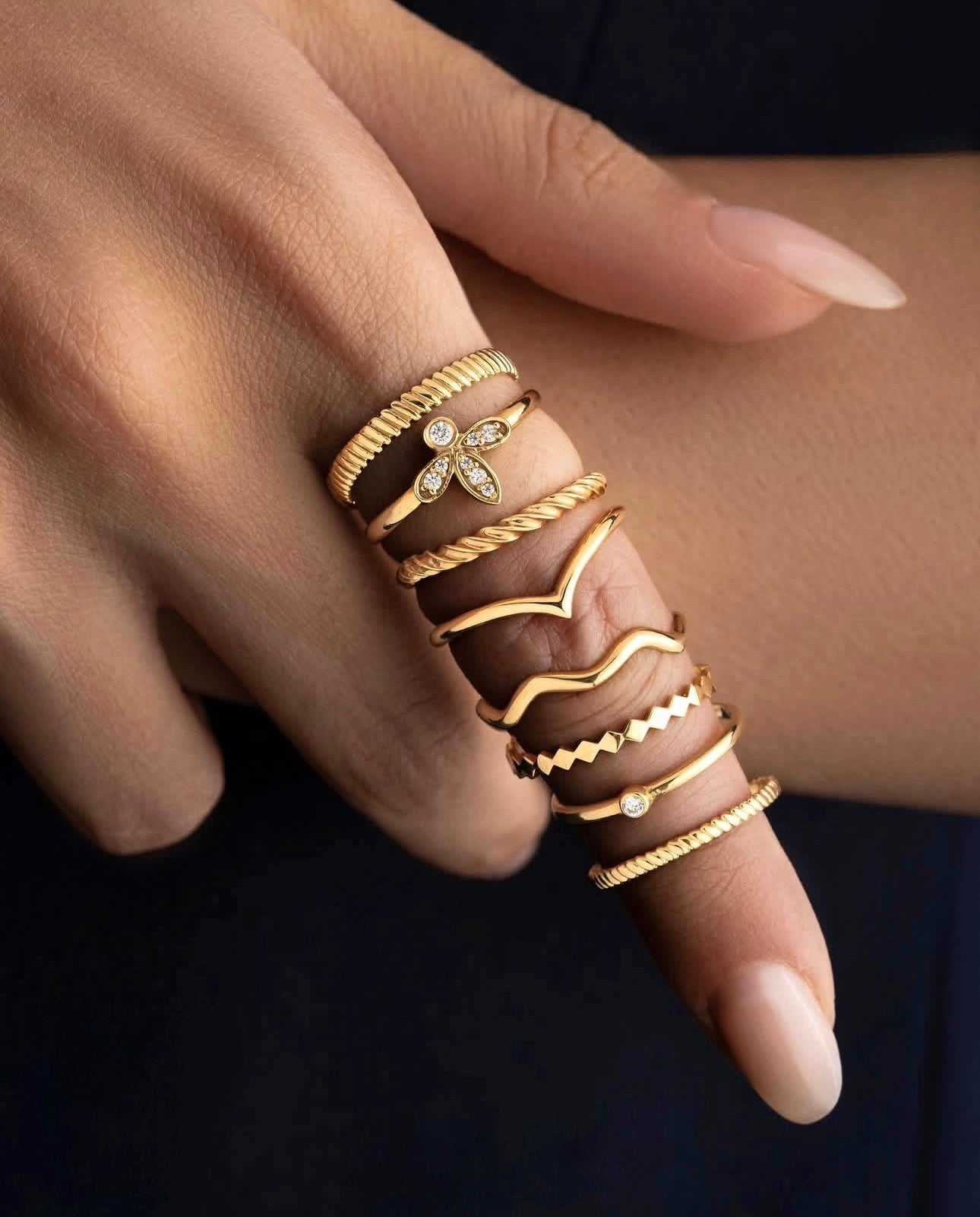 Hand mit mehreren übereinander getragenen Ringen aus 18 Karat Gelbgold, darunter der Ring „Reshhe-ye Tala“ – feines, gedrehtes Goldfaden-Design, das Eleganz und Schlichtheit in einem modernen Look verkörpert.