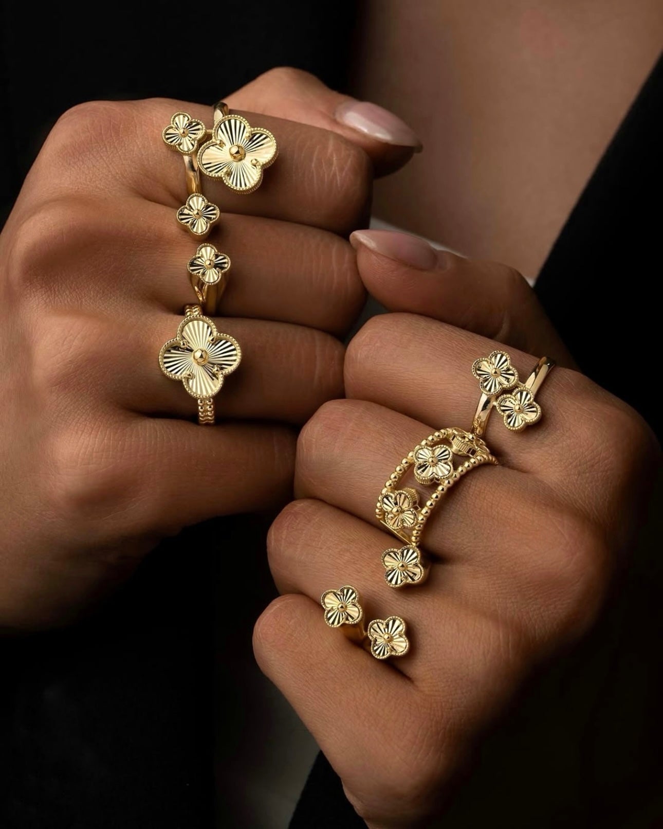 Nahaufnahme eleganter 18k-Goldringe an beiden Händen; mehrere filigrane, vierblättrige Blütenelemente mit geriffelter Struktur und polierten Goldkugeln in der Mitte; warmes Licht, schwarzer Hintergrund, luxuriöser Look.