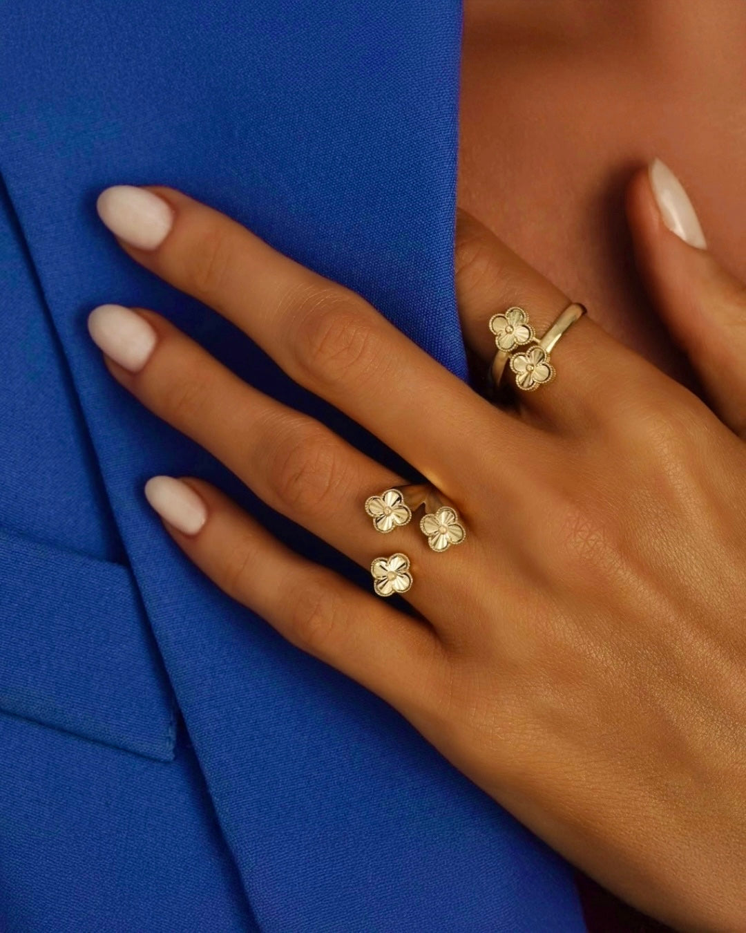 Elegante Hand mit hellen, mandelförmigen Nägeln, die einen royalblauen Blazer hält. An den Fingern mehrere goldene Ringe mit kleinen vierblättrigen Blütenelementen, stilvoll arrangiert und klar erkennbar.