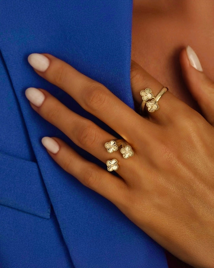 Elegante Hand mit hellen, mandelförmigen Nägeln, die einen royalblauen Blazer hält. An den Fingern mehrere goldene Ringe mit kleinen vierblättrigen Blütenelementen, stilvoll arrangiert und klar erkennbar.