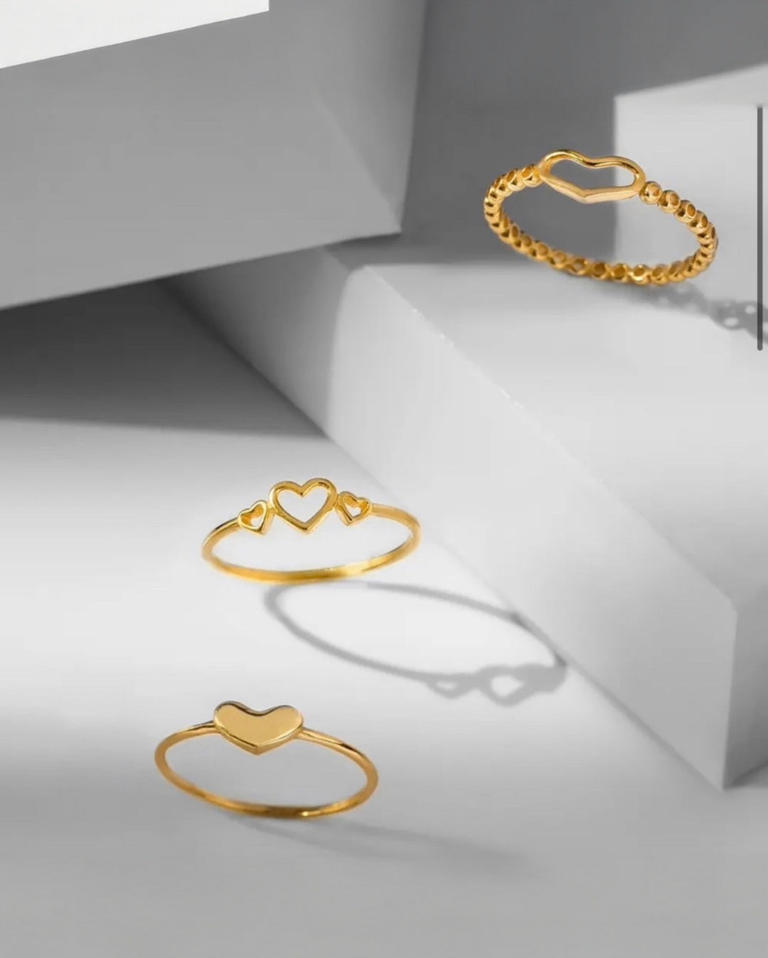 Elegante Aufnahme von drei filigranen Goldringen mit Herzmotiven auf minimalistischen, geometrischen Podesten.
Der obere Ring zeigt ein offenes Herzdesign mit Kugelstruktur, der mittlere Ring drei offene Herzen in unterschiedlicher Grösse, und der untere Ring ein schlichtes, poliertes Herz. Das warme Gold kontrastiert stilvoll mit dem kühlen grauen Hintergrund und den weichen Schattenwürfen, wodurch eine moderne, luxuriöse Atmosphäre entsteht.