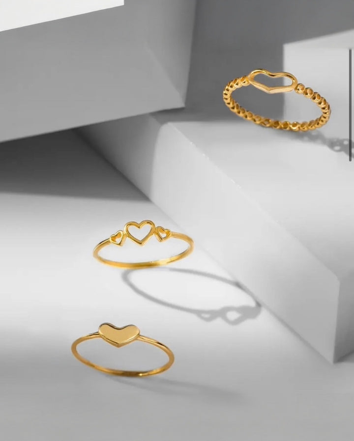 Elegante Aufnahme von drei filigranen Goldringen mit Herzmotiven auf minimalistischen, geometrischen Podesten.
Der obere Ring zeigt ein offenes Herzdesign mit Kugelstruktur, der mittlere Ring drei offene Herzen in unterschiedlicher Grösse, und der untere Ring ein schlichtes, poliertes Herz. Das warme Gold kontrastiert stilvoll mit dem kühlen grauen Hintergrund und den weichen Schattenwürfen, wodurch eine moderne, luxuriöse Atmosphäre entsteht.