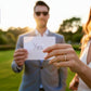 Nahaufnahme einer Frauenhand mit filigranem Diamantring bei einem Heiratsantrag im Freien, im Hintergrund ein Mann im Anzug, der ein Kärtchen mit der Aufschrift „Yes“ hält, warmes Abendlicht.