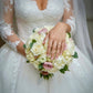 Braut in elegantem Spitzenhochzeitskleid hält einen Blumenstrauß aus weißen und zartrosa Rosen, Nahaufnahme der Hand mit feinem Diamantring, romantische Hochzeitsästhetik.